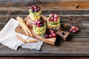 Drei Emmentaler AOP Käse Weinfässer mit Trauben und Kräutern, mit Grissinistangen auf einem rustikalen Holzbrett.